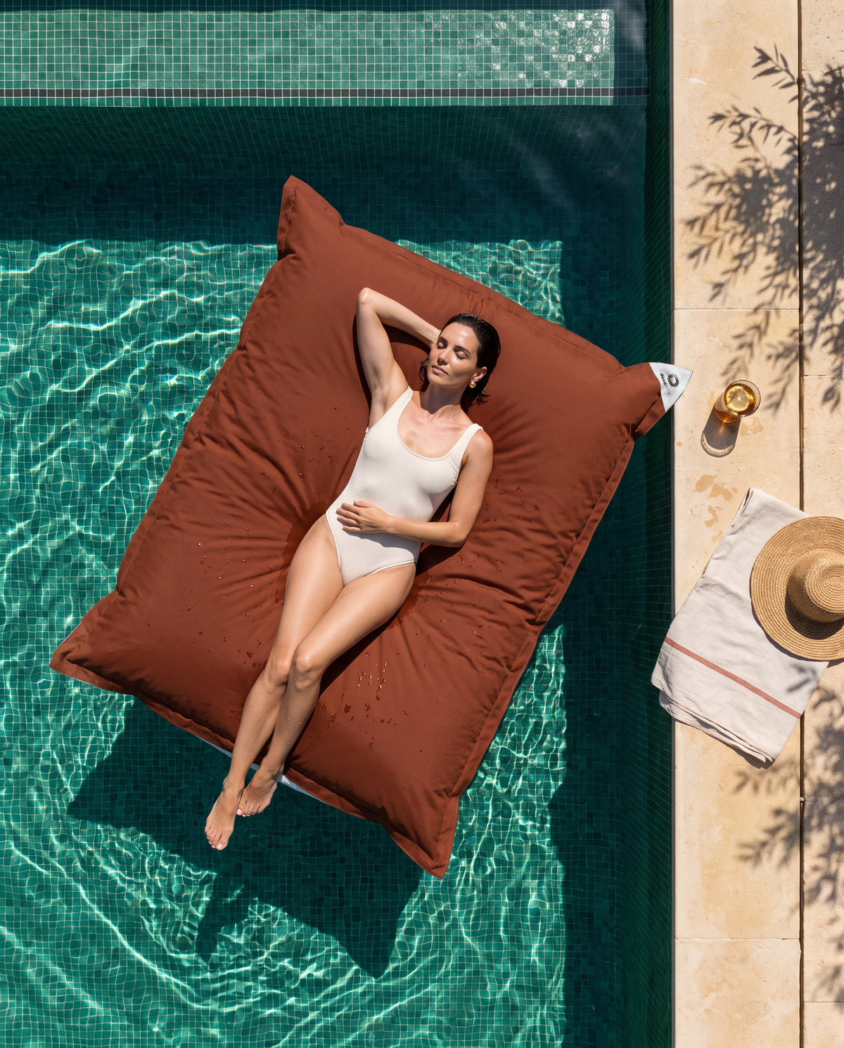 Pouf géant flottant terracotta Deco Arts sur une piscine en mosaïque verte, vue du dessus, femme allongée au soleil